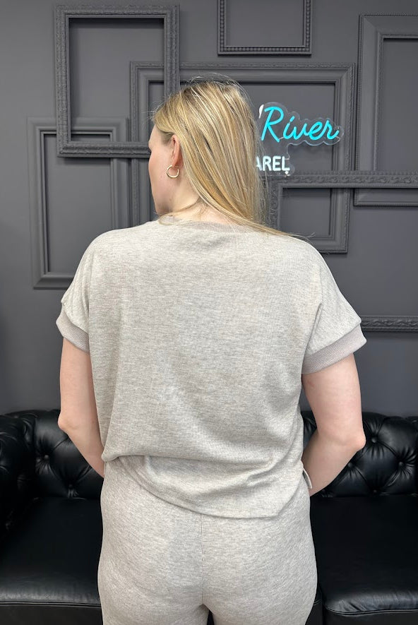 Person wearing a gray outfit standing in front of a dark wall with framed pictures and a neon sign.