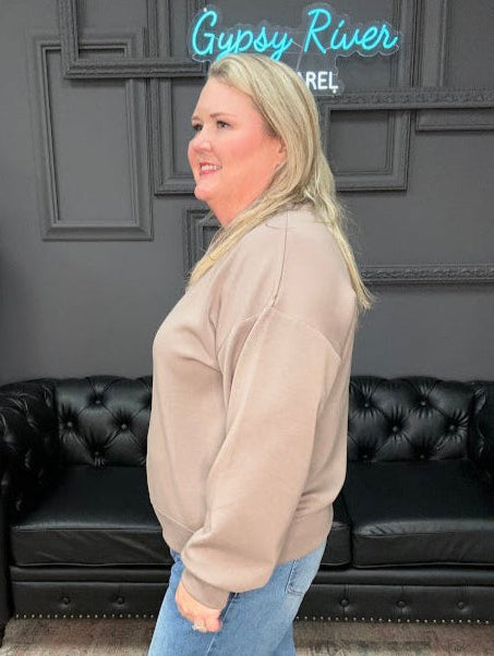 Woman in beige sweatshirt and blue jeans standing in a room with a black leather couch and decorative wall.