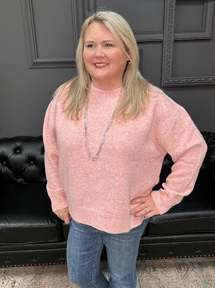 Woman wearing a pink sweater and blue jeans standing in front of a black leather couch.