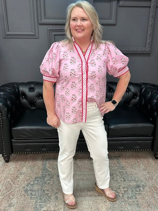 Woman wearing a pink floral blouse and white pants standing in a room with a black leather couch.