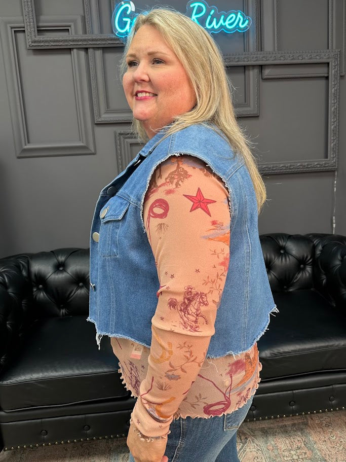 Woman wearing a denim vest over a patterned long-sleeve shirt in a room with black leather couches and a neon sign.