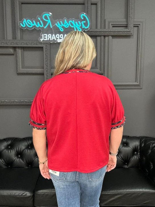 Person wearing a red top and blue jeans standing in front of a black couch with a gray wall and 'Grape Vine Apparel' sign in the background.