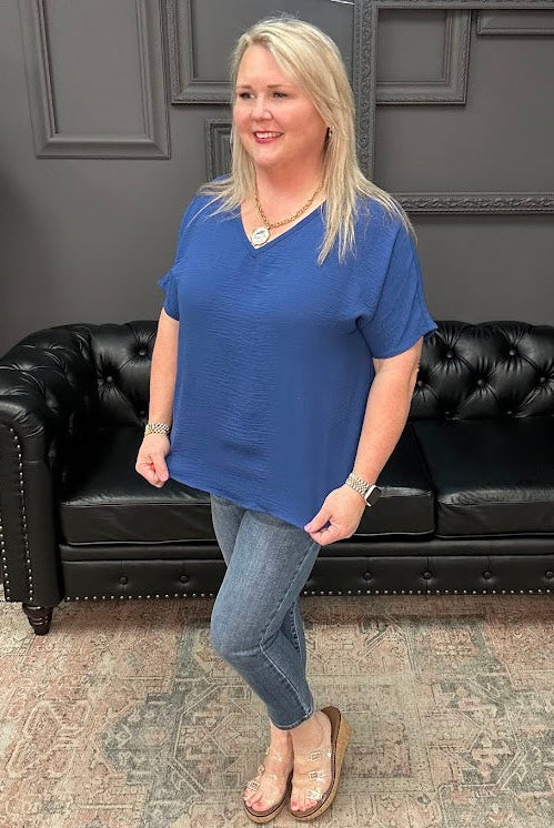 Woman in a blue shirt standing in front of a black leather couch with a gray wall and neon sign in the background.