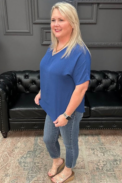 Woman in a blue shirt standing in front of a black leather couch with a gray wall and neon sign in the background.