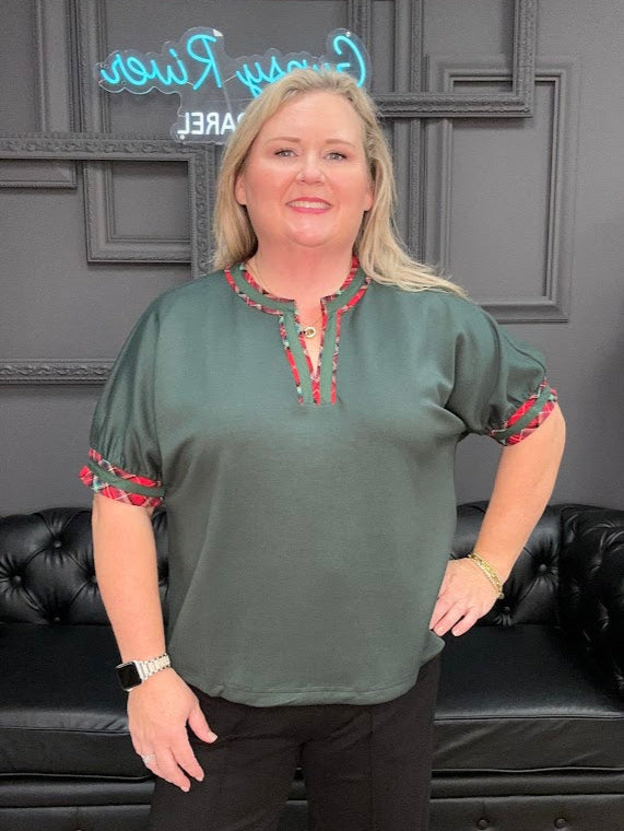 Woman wearing a green blouse with red accents in a room with black leather couches and decorative wall.