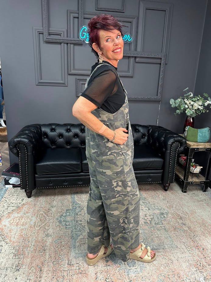 Woman wearing camouflage overalls standing in a room with a black sofa and decorative elements.