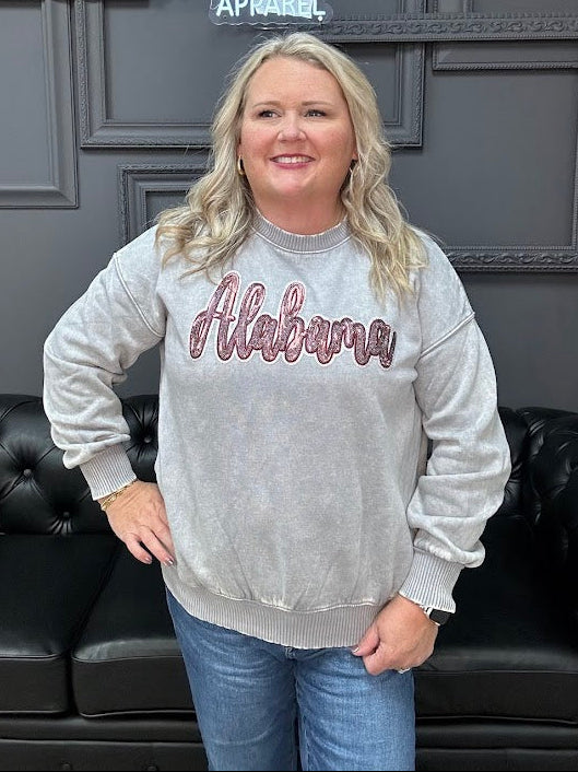Woman wearing a gray sweatshirt with 'Alabama' text in a room with a dark gray wall and black leather couch.