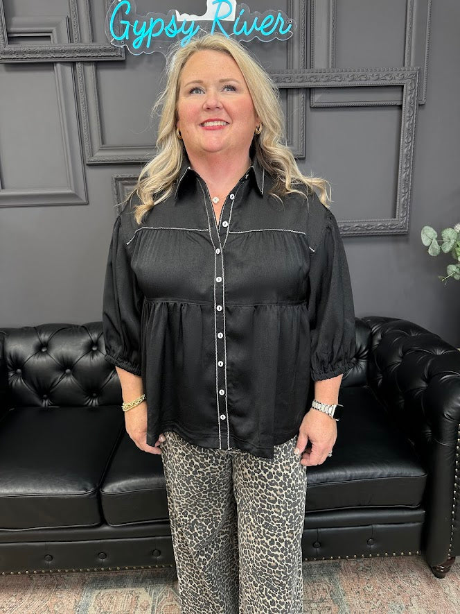 Woman wearing a black blouse and patterned pants standing in front of a black leather couch with 'Gypsy River' sign.