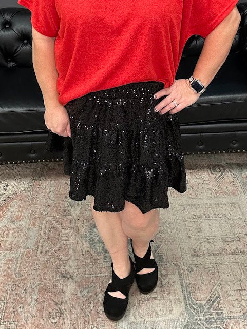Woman wearing a red top and black skirt standing in a room with a black leather couch and decorative items.