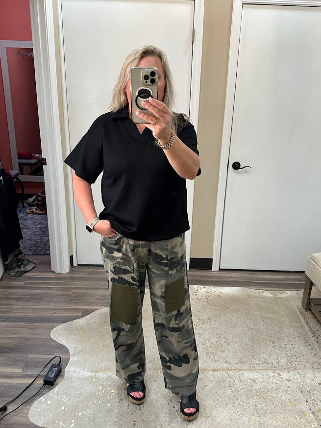 Person wearing a black polo shirt and camouflage pants taking a mirror selfie.