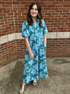 Woman wearing a teal floral dress standing against a brick wall.
