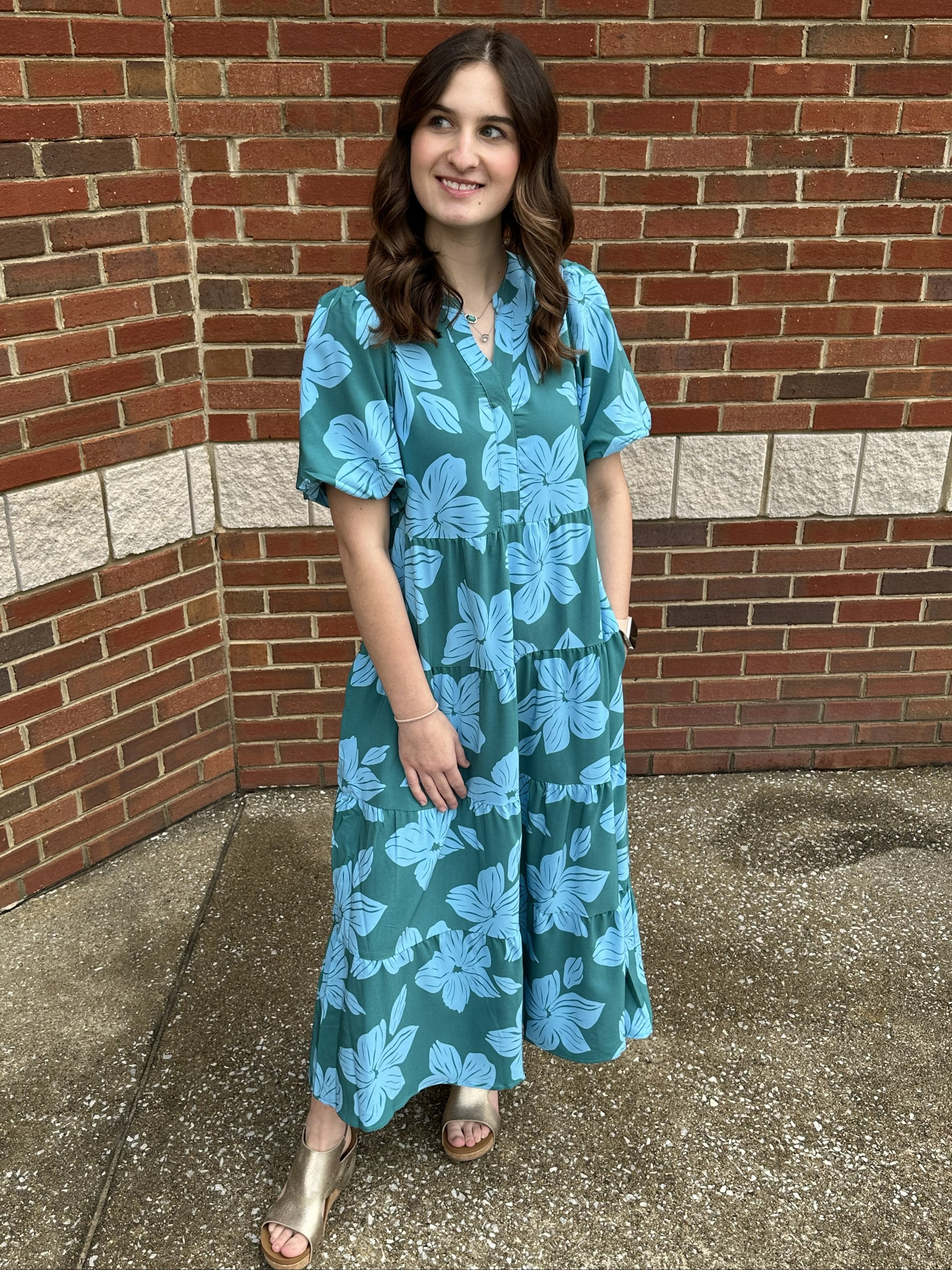 Woman wearing a teal floral dress standing against a brick wall.