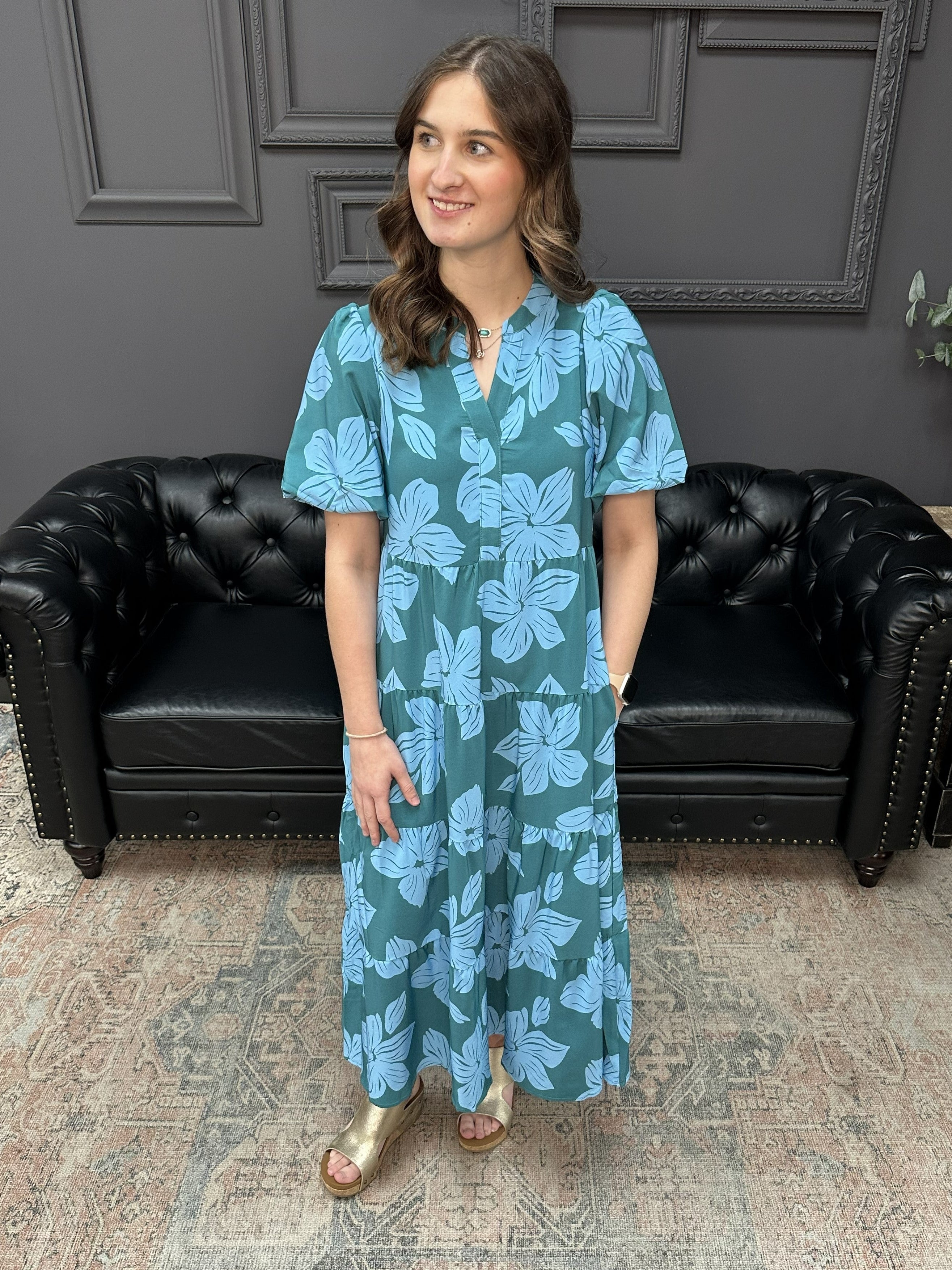Woman wearing a blue floral dress standing in a room with a black leather couch and gray walls.