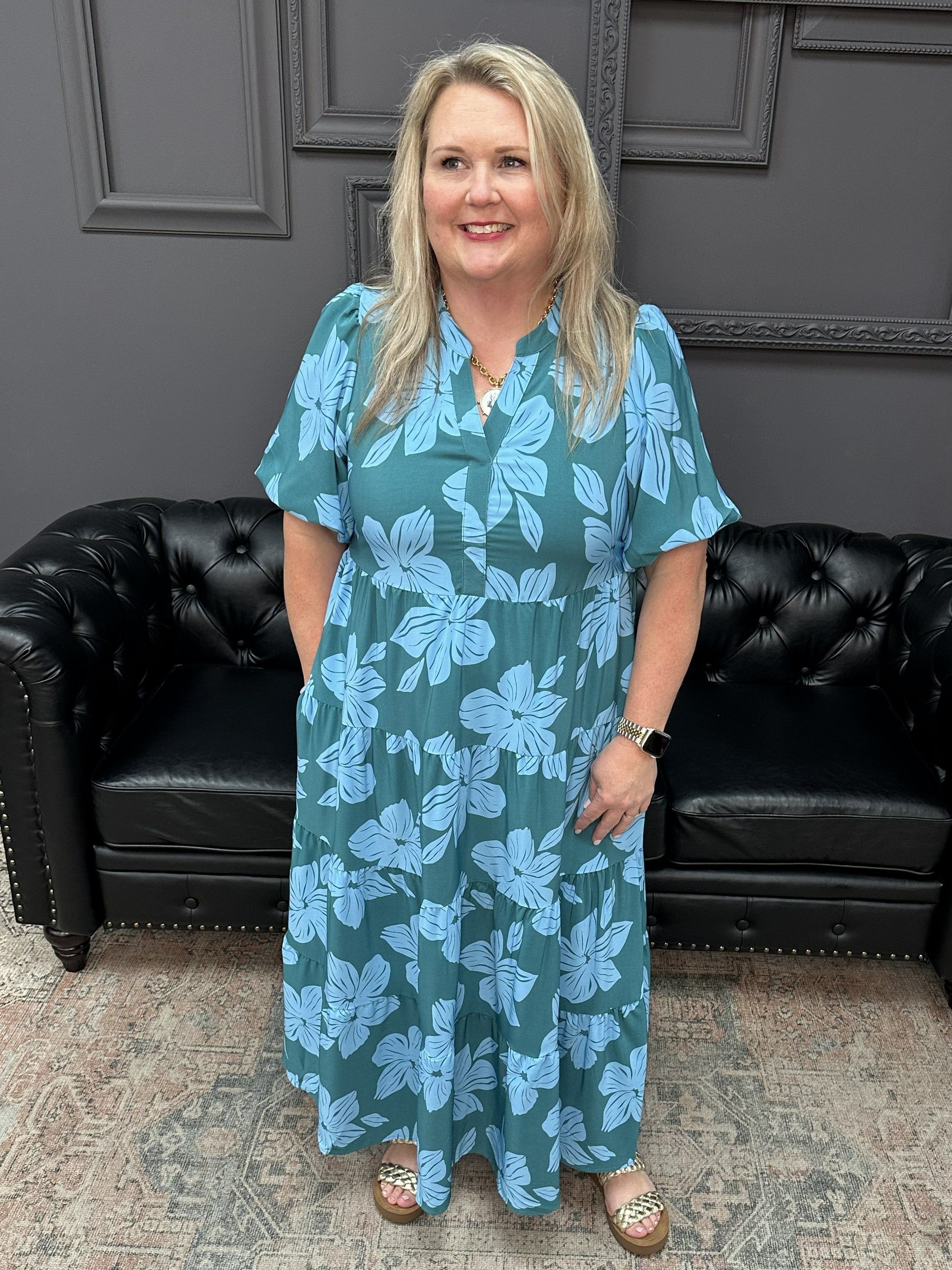 Woman in a teal floral dress standing in a room with black leather couches and a patterned rug.