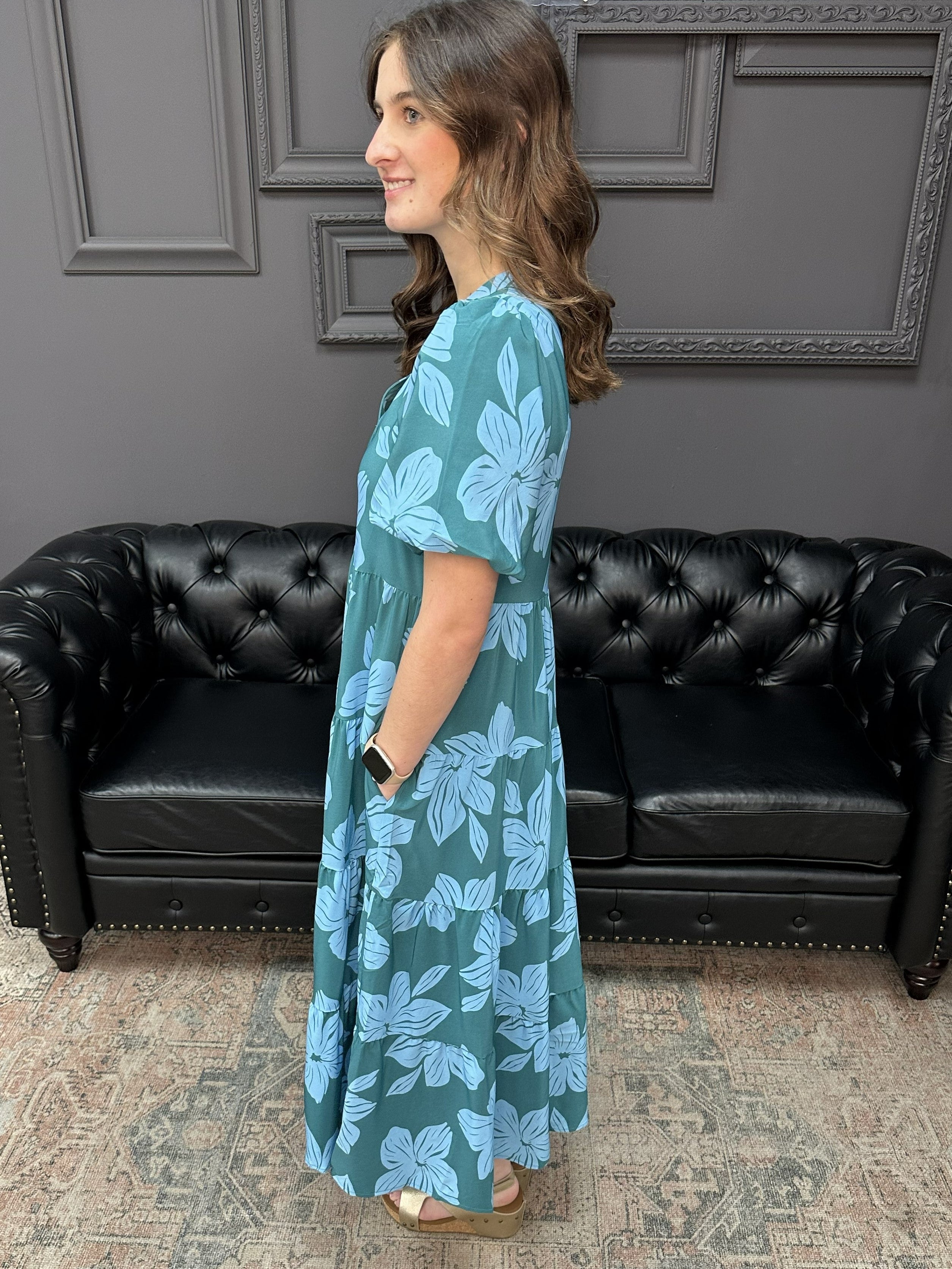 Woman wearing a blue floral dress standing in a room with a black leather couch and gray walls.