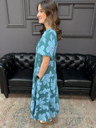Woman wearing a blue floral dress standing in a room with a black leather couch and gray walls.