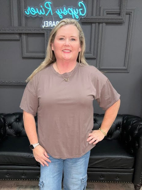 Woman wearing a brown t-shirt and blue jeans standing in a room with a black couch and decorative items.