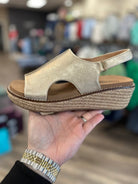 Gold wedge sandal held in a hand with a blurred store background