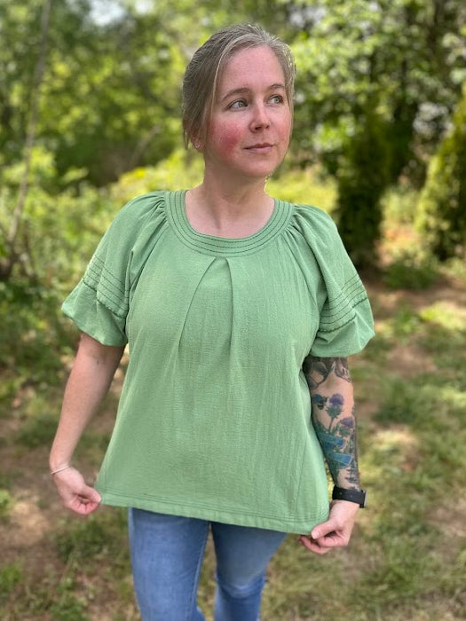Woman wearing a green blouse and blue jeans standing outdoors with trees in the background