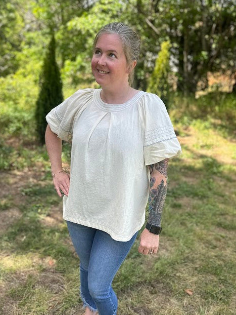 Woman standing outdoors in a natural setting wearing a light-colored blouse and blue jeans.