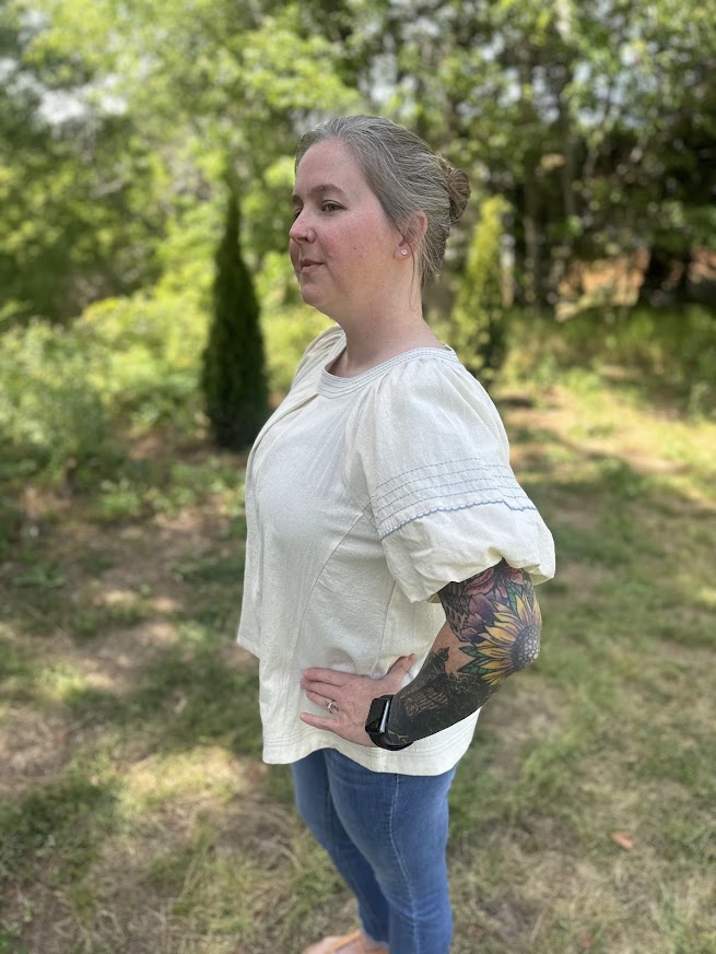 Woman wearing a white blouse and blue jeans standing outdoors with greenery in the background