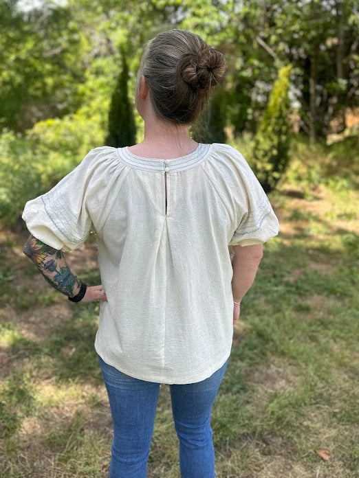 Person wearing a white blouse and blue jeans standing in a grassy area with trees.