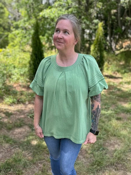 Person wearing a green shirt and blue jeans standing in a grassy outdoor area.