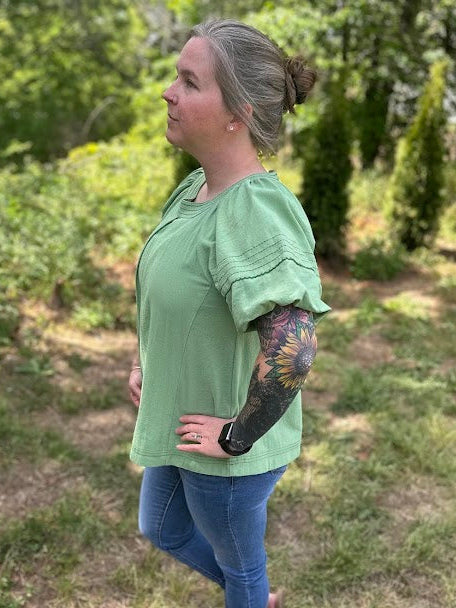 Woman wearing a green blouse and blue jeans standing in a grassy area with trees.