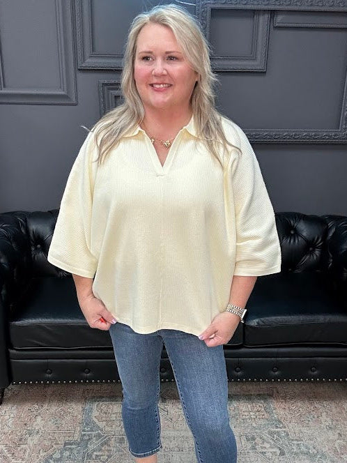 Woman wearing a light yellow blouse and blue jeans standing in a room with a black leather couch and gray walls.