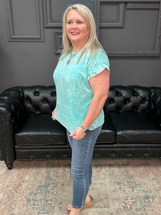 Woman in a teal tie-dye shirt and jeans standing in front of a black leather couch.