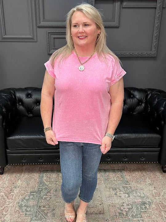 Woman in a pink shirt standing in front of a black leather couch with a gray wall and neon sign in the background.