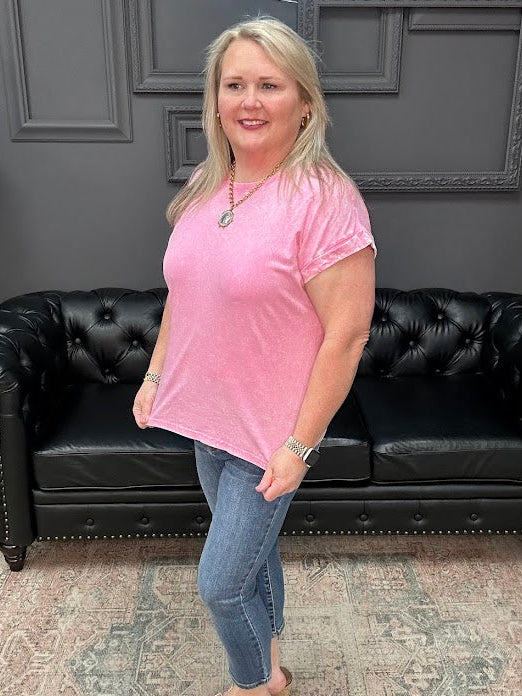 Woman in a pink shirt standing in front of a black leather couch with a gray wall and neon sign in the background.