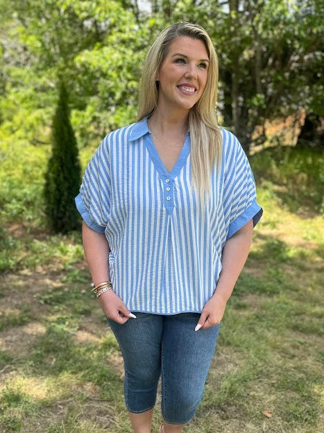 Woman wearing a blue and white striped shirt and jeans standing in a grassy area with trees.