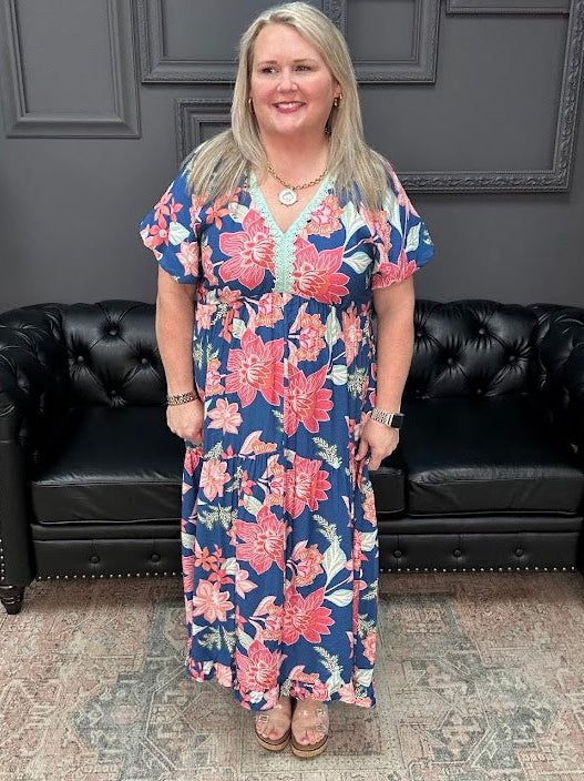 Woman in a floral dress standing in front of a black leather couch with a dark gray wall and neon sign in the background.