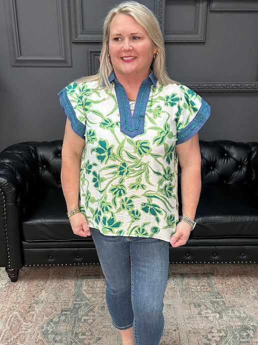 Woman wearing a patterned top and jeans standing in a room with a black leather couch and gray walls.