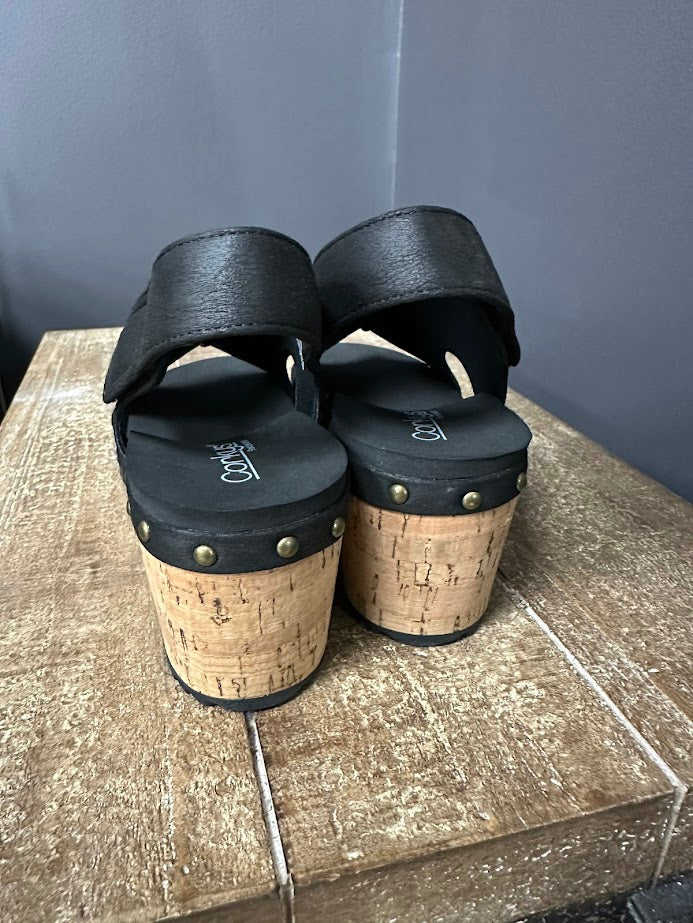 Black sandals with cork soles on a wooden surface against a gray wall.