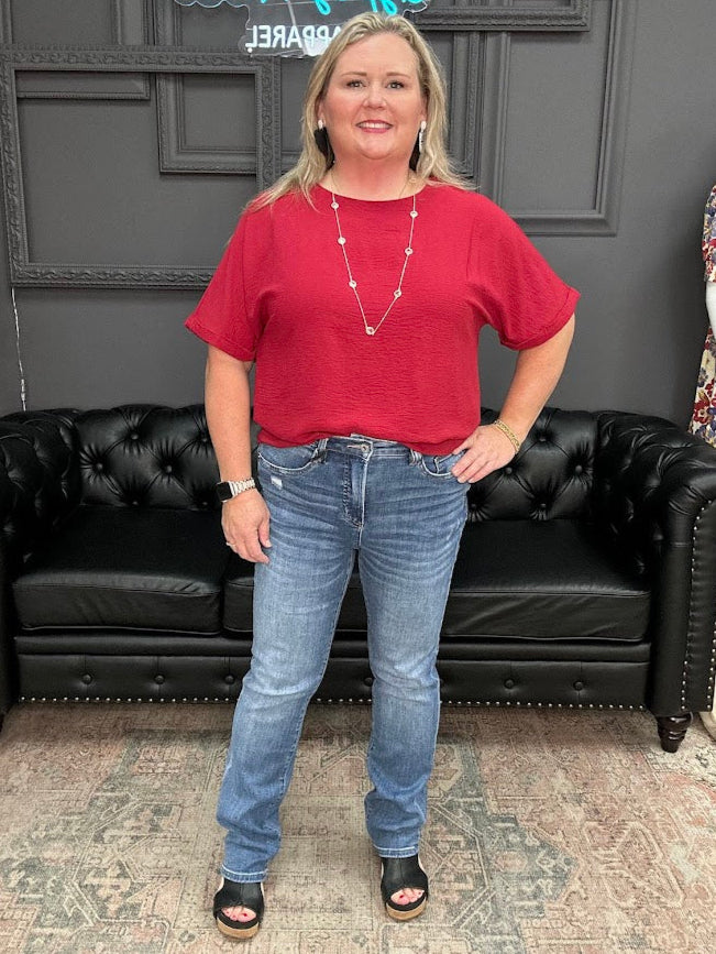 Woman wearing a red shirt and blue jeans standing in front of a black leather couch.