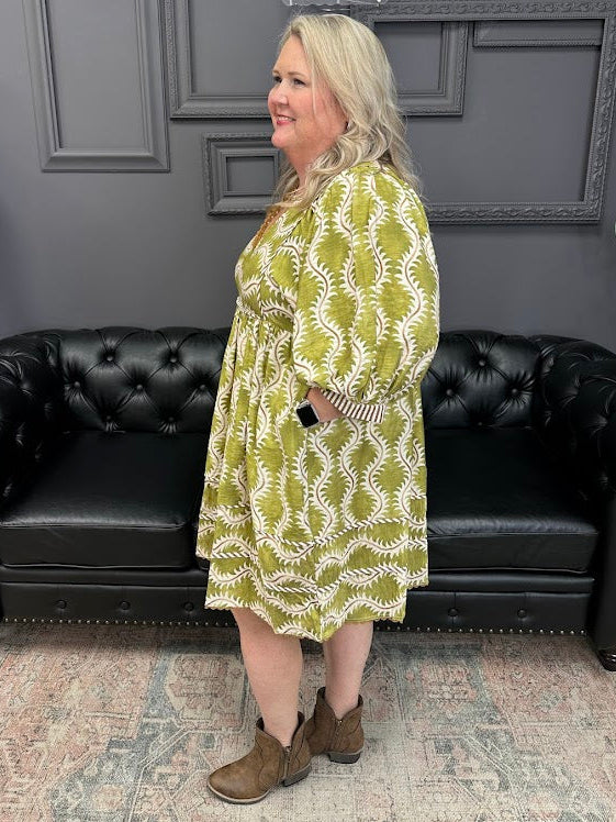 Woman wearing a green and white patterned dress standing in front of a black leather couch.