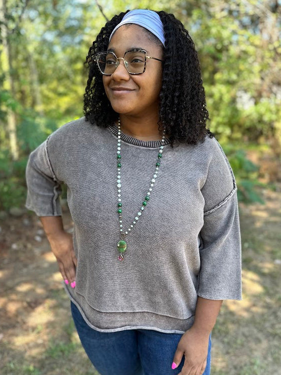 A woman wearing a gray knit crewneck sweater, blue jeans, a light blue headband, and a green beaded necklace, standing outdoors in a wooded setting.