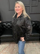 Woman wearing a black embellished jacket standing in a room with a black couch and framed pictures on the wall.