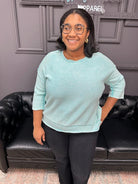 Woman wearing a light blue sweater and black pants standing in front of a black leather couch.