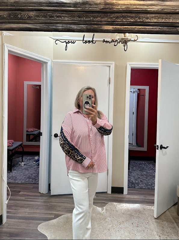 Person taking a mirror selfie wearing a pink shirt and white pants in a room with wooden flooring and a decorative mirror.