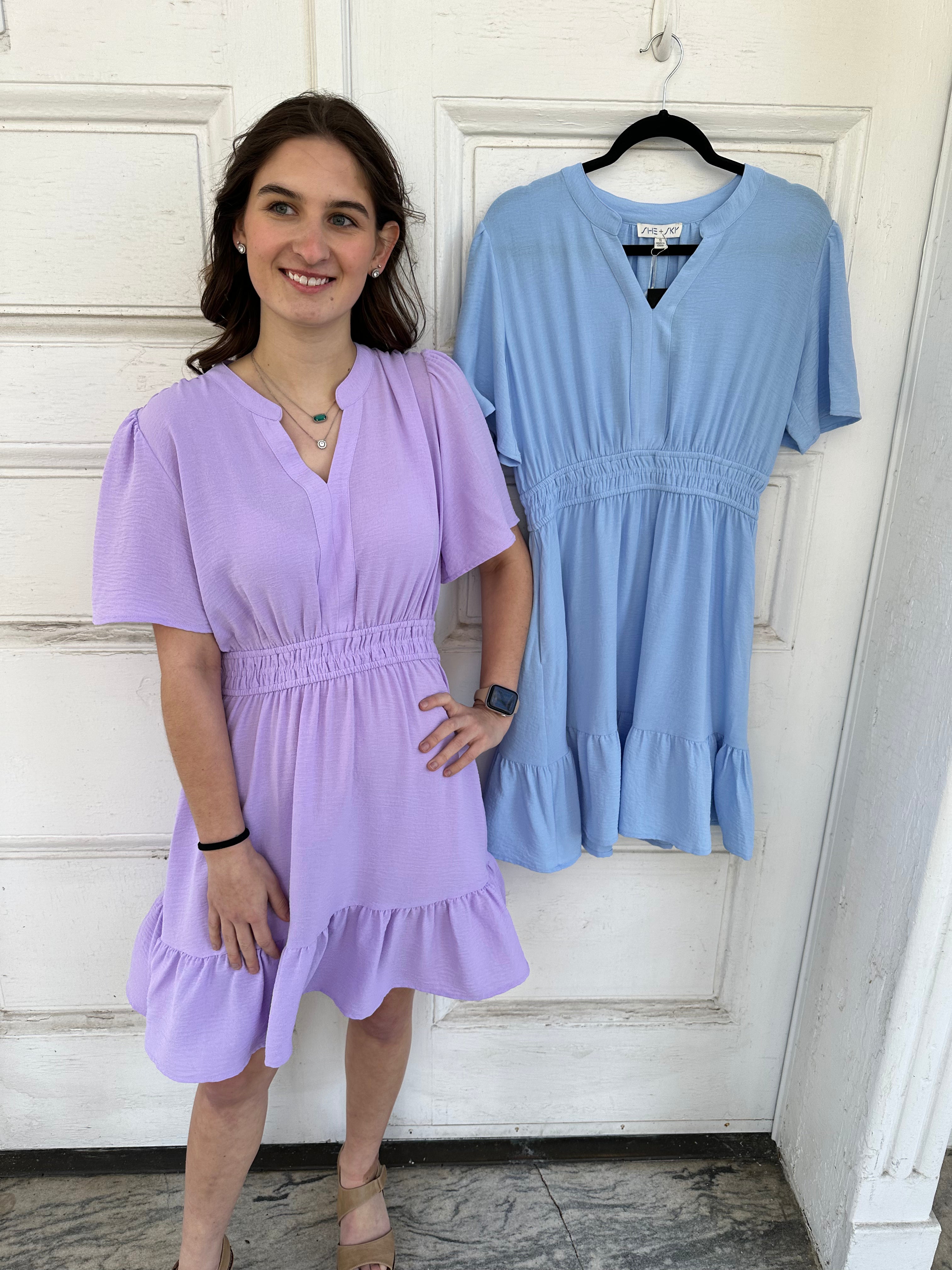 Woman wearing a purple dress standing next to a blue dress on a hanger against a white door.