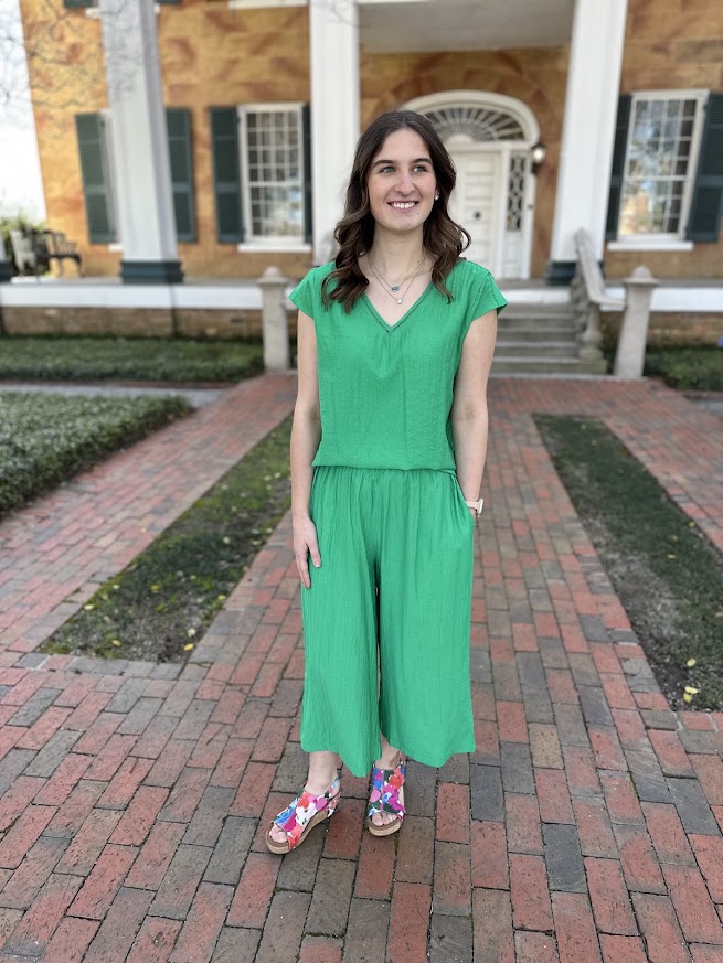 Woman in a green jumpsuit standing on a brick path in front of a building.