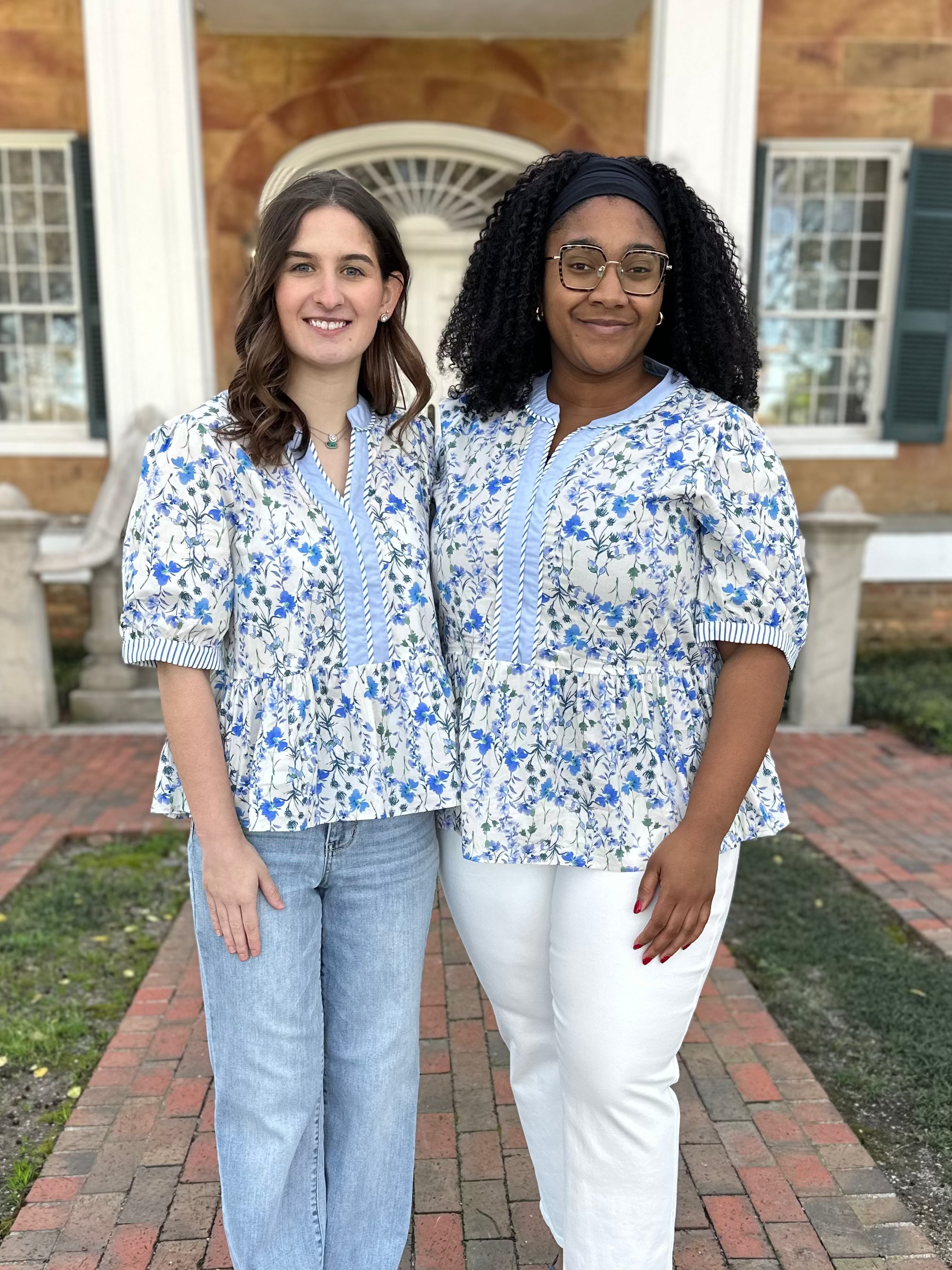 Two women in matching floral shirts standing on a brick path in front of a building.