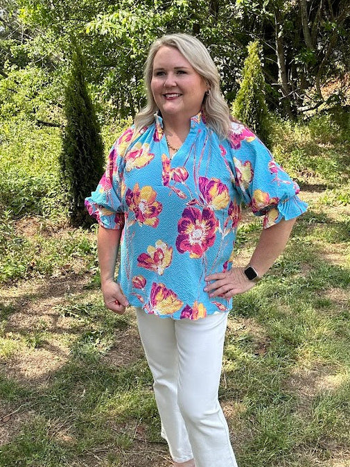 Woman wearing a colorful floral blouse and white pants standing in a grassy area with trees.
