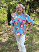 Woman wearing a colorful floral blouse and white pants standing in a grassy area with trees.
