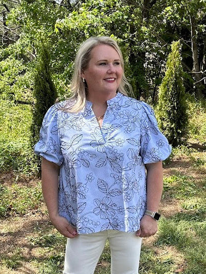 Woman standing outdoors in a blue floral blouse and white pants, surrounded by greenery.