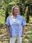 Woman standing outdoors in a blue floral blouse and white pants, surrounded by greenery.