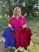 Woman holding two shirts, one blue and one red, outdoors with trees in the background.